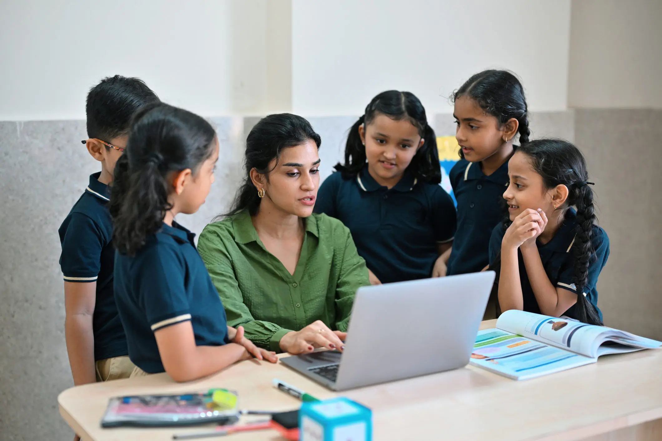 A teacher sits at a desk using a laptop while a group of cheerful primary school students gathers around her, engaged and curious. Digital learning, modern education, teacher–student interaction, classroom technology, teamwork, and collaborative learning environments.